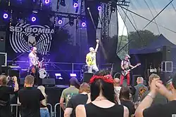 Zdjęcie przedstawiające tłum ludzi stojących na trawie przed sceną podczas festiwalu. Na scenie widać kolorowe światła oraz grupę muzyków zespołu Sexbomba. Wokalista stoi przy mikrofonie i wznosi w górę rękę.