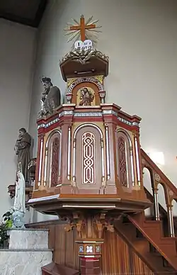 Zdobiona, drewniana neogotycka ambona z daszkiem, o podstawie w kształcie ośmiokąta, widoczna rzeźba gołębicy, tablic przykazań i promieniującego krzyża. W tle zasłonięte figury