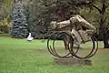 zdjęcie rzeźby plenerowej. Na betonowym postumencie umieszczone są dwa duże metalowe koła, każde składa się z trzech metalowych obręczy, przesuniętych względem siebie. Środkowa obręcz jest przerwana. Na kołach umieszczone są dwie bryły, podobne do rowerzystów, mocno pochylone do przodu. Zauważyć można głowy i torsy z zaznaczonymi ramionami. Całość umieszczona na trawniku, w tle widoczne drzewa iglaste.