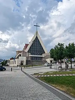 Budynek, ze ścianą frontową układającą się w trójkąt, wypełniona witrażem. na szczycie trójkąta znajduje się duży, miedziany krzyż