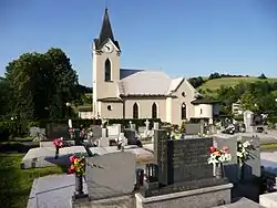 Zdjęcie przedstawia kościół sfotografowany z terenu otaczającego go cmentarza. Na pierwszym planie są współczesne nagrobki. Dalej kościół w bocznym widoku. Dominującym jego elementem jest wieża nakryta wysokim ostrosłupowym dachem. Na wieży umieszczony jest zegar. Nawa kościoła jest krótka, z dwoma wydłużonymi, ostrołukowymi oknami. W części prezbiterium znajdują się dobudówki – wysoka z trójkątnym szczytem i oknem oraz półokrągła. Za kościołem po prawej stronie są wzgórza pokryte łąkami, po lewej – grupa drzew.