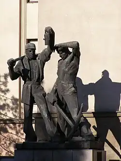 Sztygar (osoba z lewej strony) z żelazkiem (młotem) i górnik dołowy z oskardem – postaci z pomnika przed głównym budynkiem A-0 Akademii Górniczo-Hutniczej w Krakowie