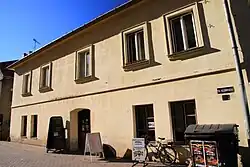 Zdjęcie przedstawia piętrowy budynek sfotografowany z ukosa. Dom pomalowany jest na żółto. Okna są prostokątne, na piętrze w profilowanych opaskach. Szeroki okap dachu rzuca cień na ścianę.