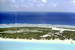 Zdjęcie. Wyspa z wysokim masztem po lewej stronie i zabudowaniami po prawej.