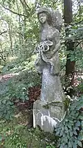Zdjęcie kamiennej rzeźby przedstawiającej smukłą kobietę w długiej sukni. Z przodu trzyma bukiet kwiatów wyglądający jak zebrane fałdy materiału. Rysy twarzy delikatne, z tyłu głowy duży kok lub kaptur. Całość w otoczeniu drzew i krzewów.