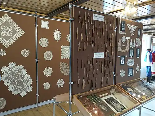 Zdjęcie fragmentu ekspozycji muzealnej, składającej się z tablicy i gabloty pośrodku pomieszczenia oraz tablicy przy ścianie. Na bliższej tablicy przymocowanych jest kilkadziesiąt par klocków o różnych kształtach i rozmiarach, opisane są jako  „Klocki z całego świata ze zbiorów Magdaleny Dyga” oraz „Klocki z Bobowej”. Obok nich wiszą wykonane techniką klockową niewielkie serwety i kołnierzyk, wraz z czarno-białymi fotografiami koronczarek w trakcie pracy. Tablica przy ścianie eksponuje również serwety z koronki klockowej, o rozmiarach znacznie większych.