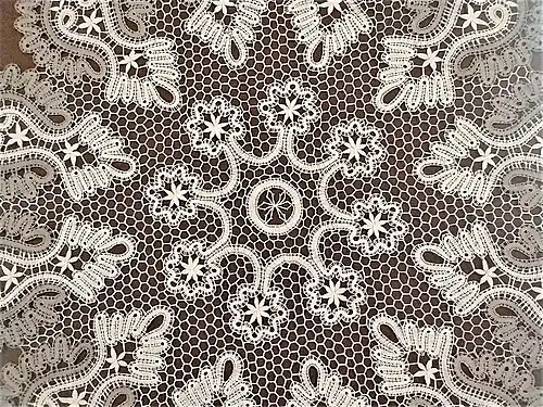 Zdjęcie centralnego fragmentu dużej serwety na planie koła, wykonanej metodą koronki klockowej. Pośrodku wyróżnia się motyw z długiego, ozdobnego paska, okalającego faliście 8 motywów z drobnych kwiatków. Obramowanie serwety składa się z symetrycznie rozłożonych, geometrycznych wzorów paskowych w dwóch kolorach: szarym oraz ecru. Części zewnętrzna i wewnętrzna połączone są drobną siatką o sześciobocznych oczkach.