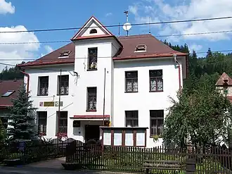 Zdjęcie przedstawia front budynku urzędu gminy w Łomnej. Budynek jest piętrowy, nakryty dachem czterospadowym. W części środkowej fasady jest ryzalit z wejściem głównym. Budynek jest w ogrodzie otoczonym drewnianym płotem. Przy płocie znajduje się ławka i tablica na ogłoszenia.