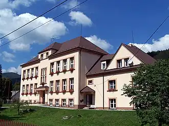 Zdjęcie wykonane w słoneczny dzień przedstawia fasadę czeskiej szkoły w Łomnej. Szkoła składa się z dwóch części, po lewej stronie jest wyższa, dwupiętrowa z dachem czterospadowym. W tej części pośrodku mieści się główne wejście do budynku. Część z prawej strony jest niższa, piętrowa, z wysokim dachem. Okna w części niższej są mniejsze, zbliżone do kwadratu. Przed budynkiem jest trawnik i drzewo. Z tyłu wyłaniają się fragmenty gór.