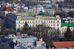  Fasada frontowa budynku Gmachu Namiestnictwa we Lwowie. Ścianę frontową zdobi szereg okien. Budynek czterokondygnacyjny. Przed wejściem do budynku znajdują się cztery kolumny. W oddali błękitne niebo.