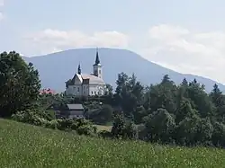 Zdjęcie przedstawia kościół w górskim krajobrazie. Na pierwszym planie jest łąka, jej kształt wskazuje, że znajduje się ona na stoku. Za nią, na wzniesieniu, znajduje się kościół, widoczny od strony prezbiterium. Kościół posiada wieżę z wysokim dachem oraz mniejszą wieżyczkę na sygnaturkę. Świątynia jest otoczona drzewami. Za kościołem w tle jest góra o łagodnie nachylonych zboczach.