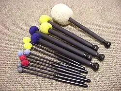 Zdjęcie przedstawia trzynaście pałek do gry na dzwonkach ręcznych. Pałki zostały ułożone obok siebie według wielkości. Pałki mają rękojeści różnej grubości oraz główki różnej wielości. Większość główek obitych jest materiałem w różnych kolorach.