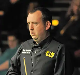 Zdjęcie. Zbliżenie na twarz łysiejącego mężczyzny spoglądającego na stół snookerowy. W dłoni trzyma kij do gry.