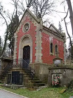 Fotografia kolorowa przedstawiająca cmentarz w pochmurny dzień. Na pierwszym planie piętrowe, neogotyckie, murowane mauzoleum ze spadzistym dachem i o czerwonych ścianach. Do głównego wejścia prowadzą schody, drzwi są metalowe. W bocznej ścianie budynku są widoczne dwa okna, a w szczytowej nad drzwiami rozeta.