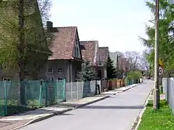 Fotografia. Asfaltowa droga biegnąca wgłąb zdjęcia, wzdłuż ulicy znajdują się identyczne, zwieńczone dwuspadowym dachem, budynki.