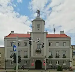 Zdjęcie przedstawia fasadę trzykondygnacyjnego budynku. Ma ona kolor szary, zaś w jej centralnej części znajduje się wieża z niewielkim balkonem, również szara. Dach obiektu jest czerwony. Na wprost wejścia do budynku widać przejście dla pieszych z sygnalizacją świetlną.