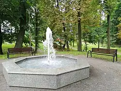 Zdjęcie przedstawia prostą fontannę koloru szarego, z której tryska woda. Obok fontanny stoją dwie ławeczki, przy jednej z nich stoi latarnia. Na drugim planie widać pozostałą, porośniętą drzewami, część parku.