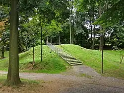 Zdjęcie przedstawia porośnięte jasnozieloną trawą wzniesienie, na którego szczycie stoją dwie ławeczki. Na szczyt pagórka prowadzą schodki ze srebrną, stalową poręczą. Przed wzniesieniem przebiega żwirowa ścieżka, zaś wokół rosną różnej wysokości drzewa.