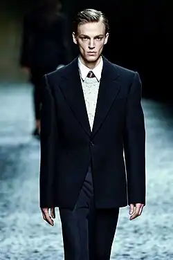 fotografia przedstawia szczupłego blondyna w czarnym garniturze i jasnej koszuli, idącego po wybiegu