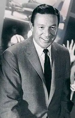 czarno-białe zdjęcie przedstawiające czarnowłosego mężczyznę w popielastym garniturze i ciemnym krawacie