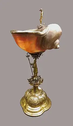 Zdjęcie przedstawia duży, dekoracyjny puchar. Czasza jest wykonana z dużej muszli, trzon i podstawa srebrne, pozłacane. Trzon to rzeźbiona postać mężczyzny z halabardą. Podstawa wypukła, pokryta rytymi ornamentami.