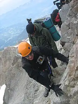 Zdjęcie. Po wąskiej grani idzie wspinacz w kasku i ciemnych okularach. W tle strome, skaliste zbocze.