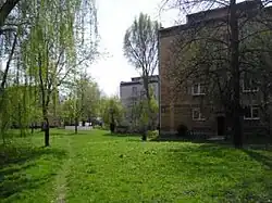 Zdjęcie. Na pierwszym planie fotografii widać zielony trawnik oraz kilka drzew. Po lewej stronie zdjęcia znajduje się kilka szarych bloków. W oddali widać więcej budynków i zieleni