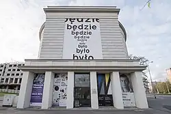 Portyk z wejściem głównym, nad nim mural „Klepsydra” Stanisława Dróżdża.