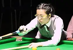 Kobieta w kamizelce w kratkę i okrągłych okularach pochylona nad stołem snookerowym. Trzyma kij do snookera przygotowując uderzenie z przyrządu.