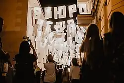 Zdjęcie zrobione po zmierzchu. Na ulicy publiczność festiwalu, ponad ich głowami białe lampiony w kształcie liter z różnych alfabetów. Instalacja artystyczna wypełnia całą szerokość ulicy.