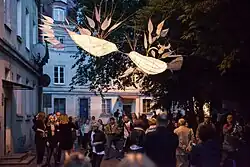 Zdjęcie wykonane po zmroku. Ulica wypełniona publicznością festiwalu, ponad głowami wiszą białe lampiony w kształcie ptaków i ryb wykonane z papieru i wikliny.