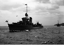 Widok od przodu z lewej ćwiartki na wolno płynący okręt, za jego rufą w tle kolejne okręty Dywizjonu Niszczycieli. Na maszcie powiewa proporzec dowódcy okrętu, na wytyku dziobowym proporzec Marynarki Wojennej. Na pokładzie dziobowym stoją w dwuszeregu marynarze. Na mostku wachta, nad nim leci mewa. Powierchnia morza lekko pofalowana, niebo zachmurzone, zza chmur, z tyłu okrętu przebija słońce.