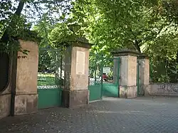 Zdjęcie przedstawia bramę wejściową parku. Brama składa się z 4 wysokich słupów, pomiędzy którymi znajdują się zielone bramki. Przed bramą znajduje się wybrukowany kostką plac.