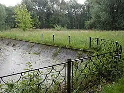 Zdjęcie z 2013 roku. Przedstawia ono zaniedbany parkowy staw ze spuszczoną wodą. Brzegi i dno zbiornika są wyłożone betonem, w którego szparach zaczynają rosnąc chwasty. Okolica stawu jest zapuszczona - przy brzegach zbiornika widać zardzewiałe ogrodzenie i pokrzywy, a dalej nieskoszoną trawę. W oddali widać drzewa i krzaki.