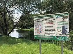 Tablica informacyjna zespołu przyrodniczo-krajobrazowego, na której uwidoczniono położenie stawu. W tle sam staw widziany od północy