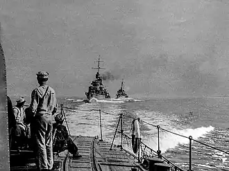 Zdjęcie z rufy „Błyskawicy” w kierunku tylnym (za rufę), na pierwszym planie fragment pokładu rufowego „Błyskawicy” z marynarzami patrzącymi w tył na pozostałe okręty Dywizjonu: w środku zdjęcia „Grom” ok. 400 m za rufa „Błyskawicy” lekko na prawo od jej śladu torowego, daleko w tle za nim i dalej na prawo sylwetka „Burzy”