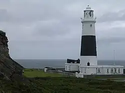 Zdjęcie. Przedstawia ono latarnię morską na Alderney. Na pierwszym planie zdjęcia widać wysoką, pomalowaną w czarne i białe pasy latarnię morską. Wieża latarni zwęża się ku górze. Na szczycie budynku jest balkon. W bryle latarni widać kilka okien. U stóp latarni widać niewielki budynek pomalowany na biało. Po lewej stronie zdjęcia można zobaczyć skały. Otoczenie latarni porośnięte jest trawą. Za latarnią, w tle widać morze.