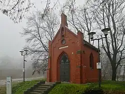 Zdjęcie przedstawia kaplicę na niewielkim wzniesieniu, w otoczeniu drzew. Widoczne są dwie ściany budynku: przednia (fasada) z wejściem w postaci drzwi koloru szarego oraz boczna z oknem. Budynek jest cały wykonany z cegły. Do kaplicy prowadzi siedem betonowych schodków, obok których stoi tablica informacyjna. Przed obiektem znajdują się dwie latarnie, zaś po jego prawej stronie widać dwie ławeczki. Zdjęcie zostało wykonane w połowie grudnia, trawa jest przyschnięta, gdzieniegdzie zalegają resztki śniegu, drzewa są pozbawione liści, a w tle widoczna jest gęsta mgła.