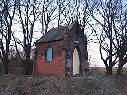 Zdjęcie przedstawia kaplicę na niewielkim wzniesieniu, w otoczeniu drzew. Widoczne są dwie ściany kaplicy: przednia (fasada) z wejściem i boczna z oknem. Zarówno wejście, jak i okno są zamurowane lub zasłonięte. Kaplica jest cała z przybrudzonej cegły, są na niej ślady przemurowań. Zdjęcie zostało wykonane wiosną lub jesienią, trawa jest przyschnięta, a drzewa pozbawione liści.