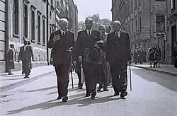 Stare, czarno-białe zdjęcie przedstawiające trzech elegancko ubranych mężczyzn idących ulicą