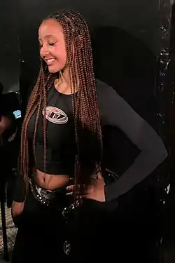 PinkPantheress ubrana w czarną, krótką bluzkę i czarne spodnie stoi uśmiechnięta z lewą ręką opartą na biodrze.