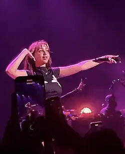 PinkPantheress ubrana w czarną bluzkę z nadrukiem, występując na scenie wyciąga mikrofon w stronę widowni i przykłada rękę do prawego ucha nasłuchując widowni.