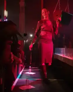 PinkPantheress podczas występu na żywo w Londynie w 2021 roku, artystka ubrana w ciemną bluzkę, spódnicę oraz wysokie buty, tanecznym krokiem porusza się na scenie, w ręku trzymając mikrofon.