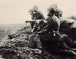 Grupa piechoty leży lub klęczy w płytkim okopie. Na pierwszym planie trójosobowa obsługa karabinu maszynowego, za nimi widocznych dwóch oficerów klęczących o krok przed okopem, widocznych także kilka karabinów wycelowanych z okopu w stronę pola widocznego po lewej stronie kadru. Trzymających je żołnierzy zasłania obsługa kaemu na pierwszym planie.