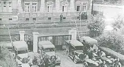 Zdjęcie czarnobiałe wykonane z okna nieznanego budynku, z wysokości ok. trzeciego piętra. Na bliższym planie grupa źołnierzy w mundurach 10 Brygady Kawalerii, dwa motocykle Sokół z koszami przykrytymi brezentem, oraz cztery półciężarówki stojące frontem do budynku. Za nimi ogrodzenie budowli wraz z zamkniętą bramą. Równolegle do niego widoczna wąska ulica po obu stronach obramowana chodnikami. Po drugiej stronie ulicy widoczne fragmenty dwóch kamienic (widoczne okna parteru) oraz grupa osób w cywilnych ubraniach stojąca na chodniku.