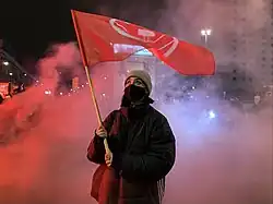 Kobieta ubrana na czarno, stojąca wśród czerwonego dymu na środku skrzyżowania, trzymająca czerwoną flagę z białą grafiką.