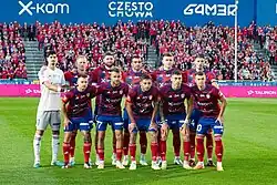 Drużyna Rakowa Częstochowa ustawiona w dwóch rzędach jeden po drugim przed domowym meczem z Arisem Limassol. Zawodnicy gospodarzy ubrani są w czerwono-granatowe trykoty meczowe, bramkarz ma na sobie biały strój.