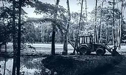 Biało-czarne zdjęcie z 2016 roku. Widać na nim prace rewitalizacyjne w parku. Na pierwszym planie widać koparkę i drzewa, po lewej stronie niewielką sadzawkę, a w tle widoczny jest nowo wybudowany, parkowy staw.