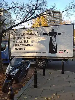 Plakat umieszczony na plandece furgonetki, na białym tle znajduje się napis: Pedofile grasują najczęściej w kościele i tradycyjnej rodzinie*. Obok znajduje się rysunek chłopca wiszącego w samych slipkach w pozie ukrzyżowania na odwróconej plecami sylwetce kapłana w czarnej sutannie. Kapłan stoi tyłem ręce ma wyprostowane i rozłożone szeroko na boki tak, że przypomina kształtem krzyż. Poniżej z prawej znajduje się logo w postaci pochylonego krzyża łacińskiego w który uderza czerwona błyskawica i tekst: Same plusy. Na dole pod głównym tekstem znajduje się linijka tekstu pisana małymi literami zaczynająca się od gwiazdki: według zgłoszeń rozpatrzonych przez Państwową Komisję ds. Pedofilii