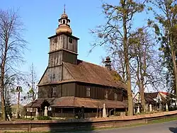 Zdjęcie wykonane w słoneczny dzień przedstawia kościół drewniany w Siedliszczach sfotografowany z drogi. Widoczne są ściana frontowa i boczna kościoła. Budynek otoczony jest sobotami, posiada wysoki dwuspadowy dach. Nad wejściem znajduje się masywna, wtopiona w bryłę budynku wieża, nakryta hełmem. Nad prezbiterium w dachu znajduje się niewielka wieżyczka na sygnaturkę. Kościół otoczony jest cmentarzem, a od drogi ogrodzony niskim drewnianym ogrodzeniem. W otoczeniu kościoła rosną drzewa, na zdjęciu - bez liści. Po prawej stronie, za kościołem, znajdują się parterowe domy mieszkalne.